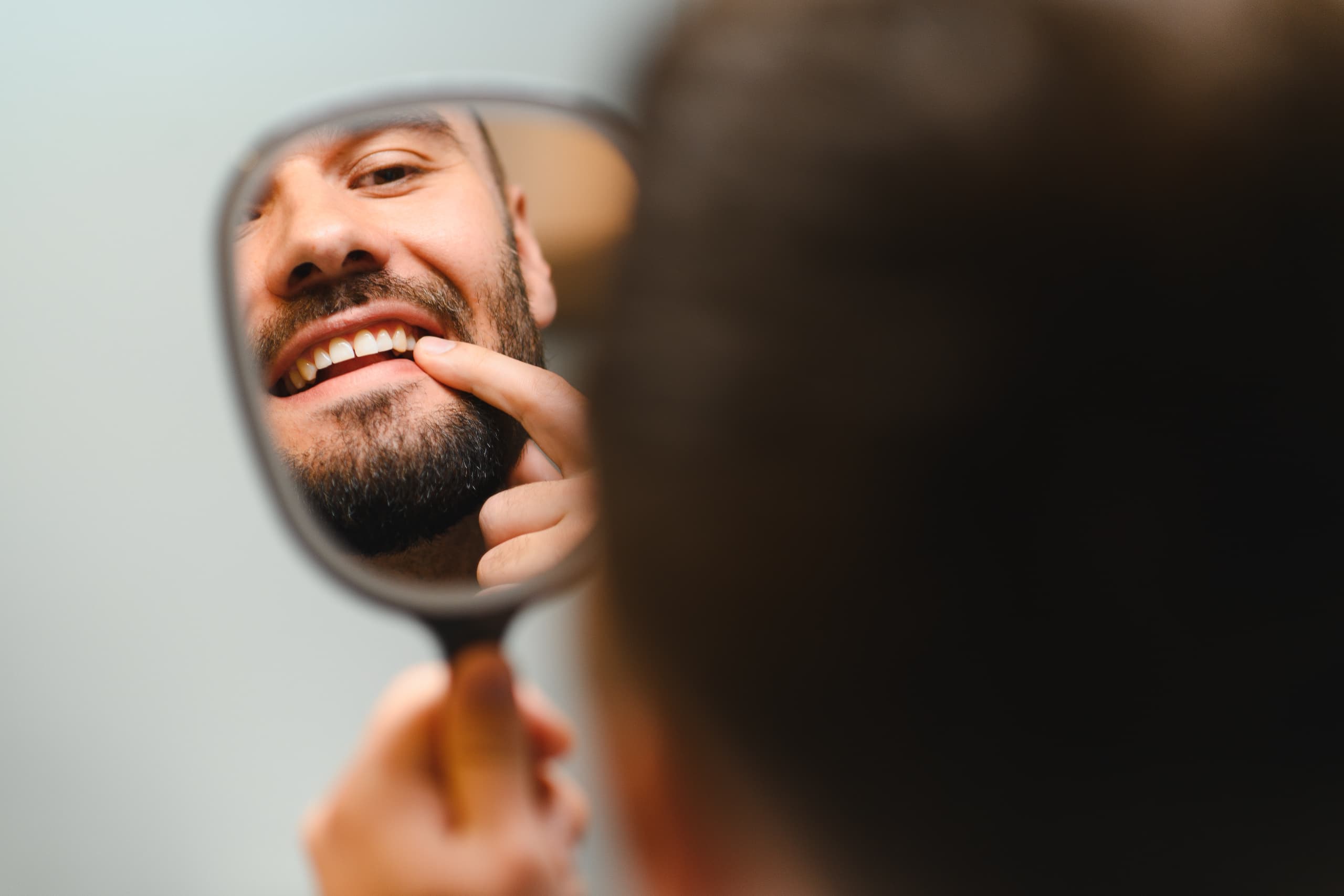 A man smiling and checking his teeth closely in a handheld mirror.