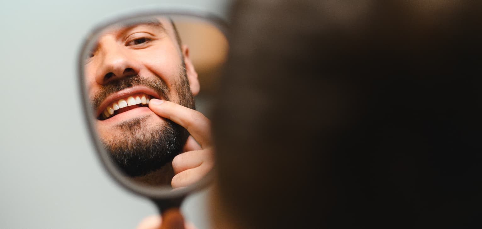 A man smiling and checking his teeth closely in a handheld mirror.